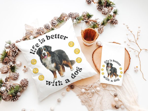 May include: White pillow and towel set with a Bernese Mountain Dog illustration and the text "life is better with a dog." Yellow tennis balls are also featured in the design. A wooden bucket and decorative garland are in the background.